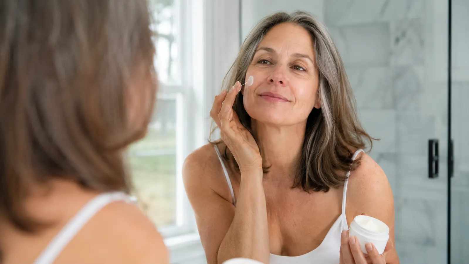 Woman applying gel cream to her face in front of a bathroom mirror, morning routine